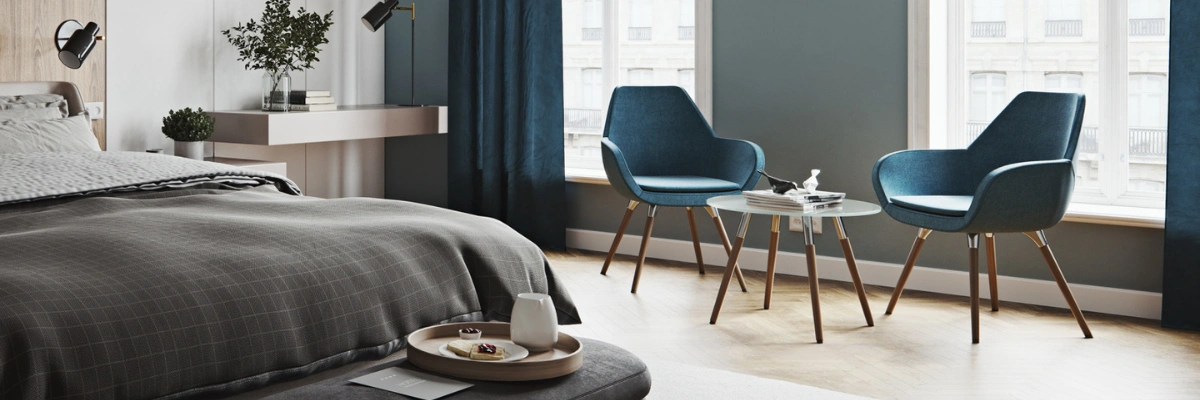Dwa morskoniebieskie fotele Profim Fan 10HW na drewnianych nogach ustawione przy okrągłym stoliku w eleganckim, jasnym apartamencie hotelowym.